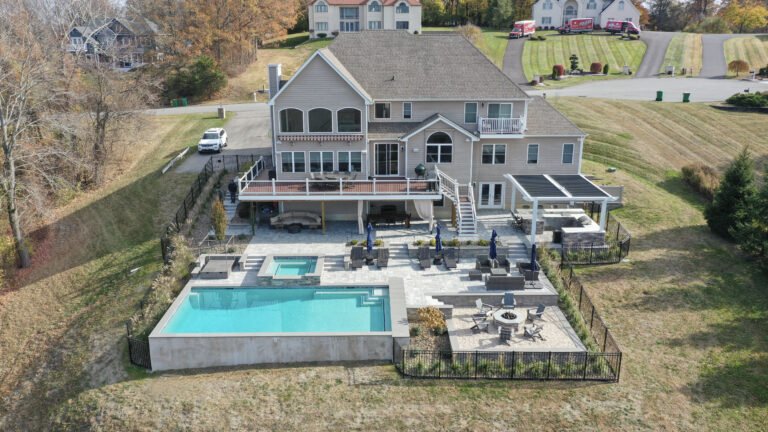 A stunning aerial view of a luxury backyard in the Hudson Valley featuring a multi-level outdoor living space with a deck, covered patio, Pergola X, a large pool with a hot tub, and a fire pit area surrounded by beautiful fall foliage