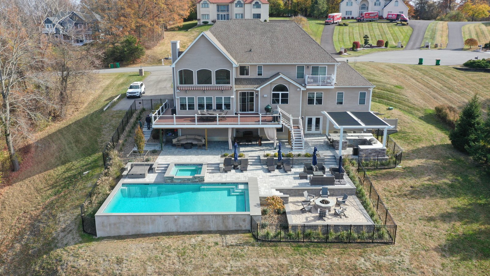 A stunning aerial view of a luxury backyard in the Hudson Valley featuring a multi-level outdoor living space with a deck, covered patio, Pergola X, a large pool with a hot tub, and a fire pit area surrounded by beautiful fall foliage